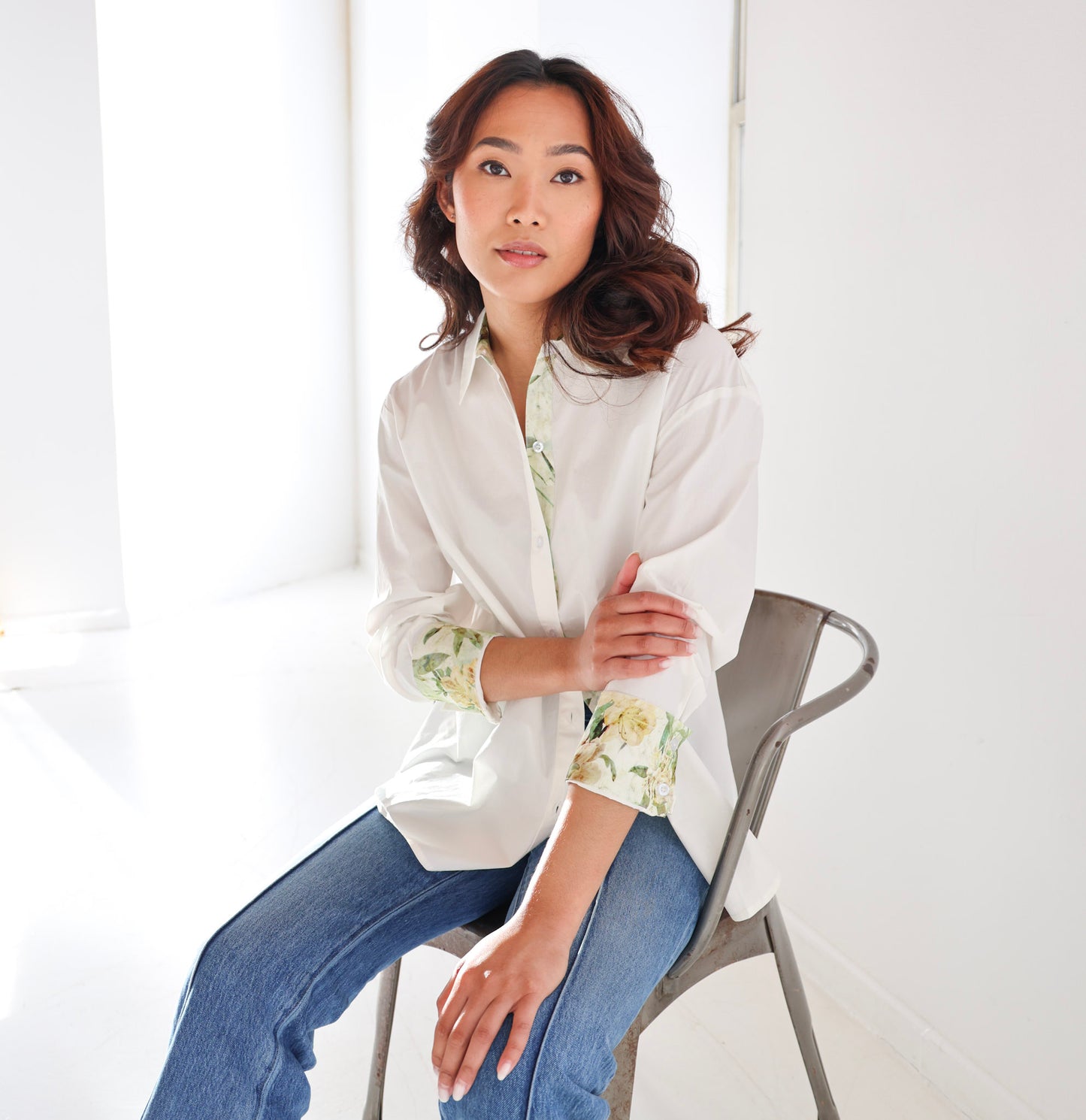 Woman sitting on a chair wearing a white blouse with floral cuffs and blue jeans.