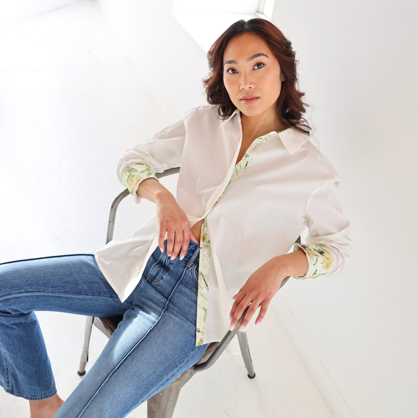 Woman sitting on a chair wearing a white blouse with floral cuffs and blue jeans against a white background