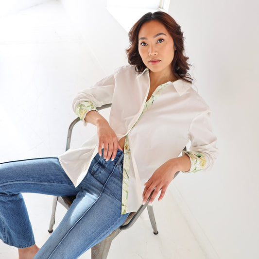 Woman sitting on a chair wearing a white blouse with floral cuffs and blue jeans against a white background