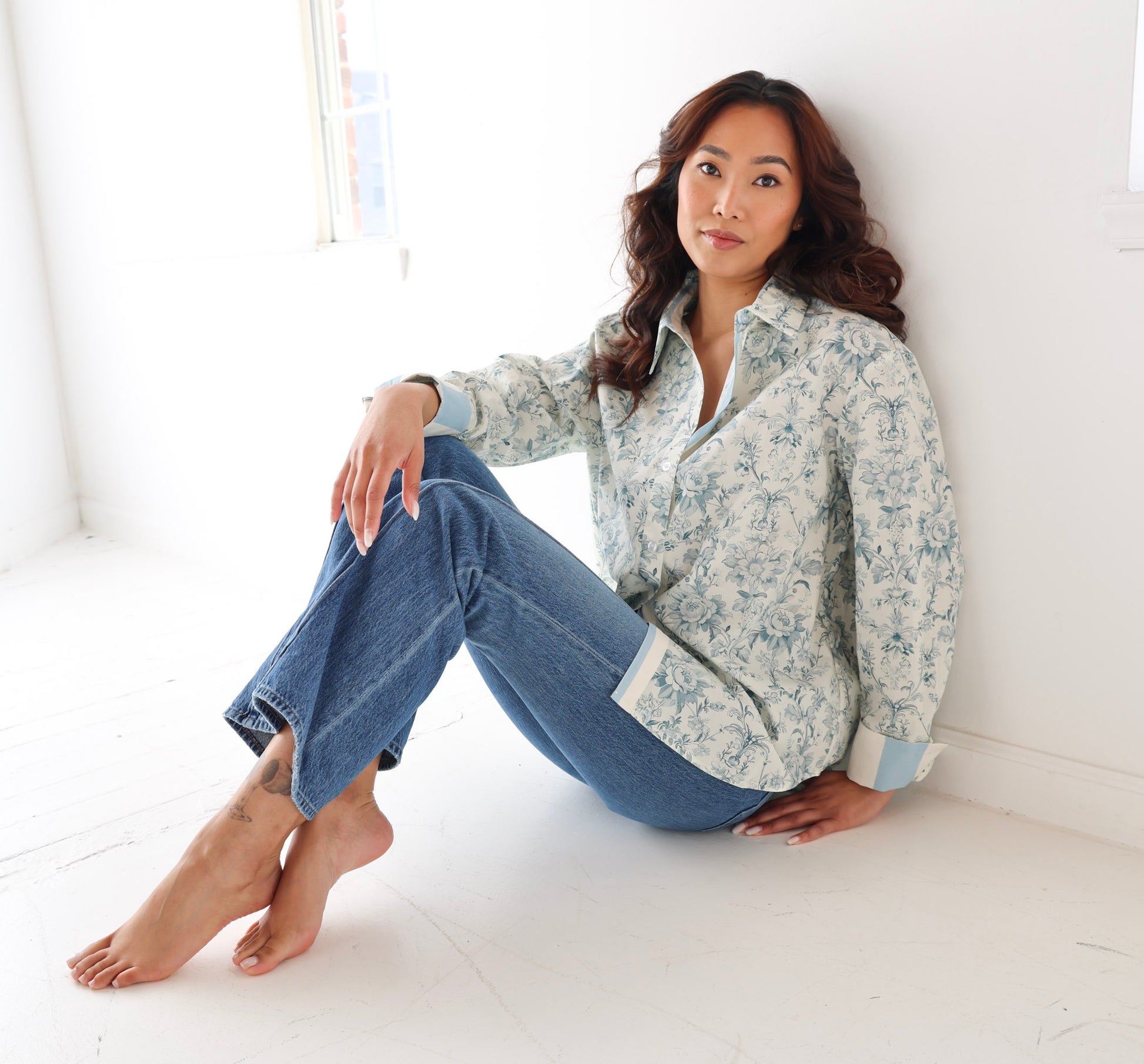Woman sitting on a white floor wearing a floral shirt and blue jeans.