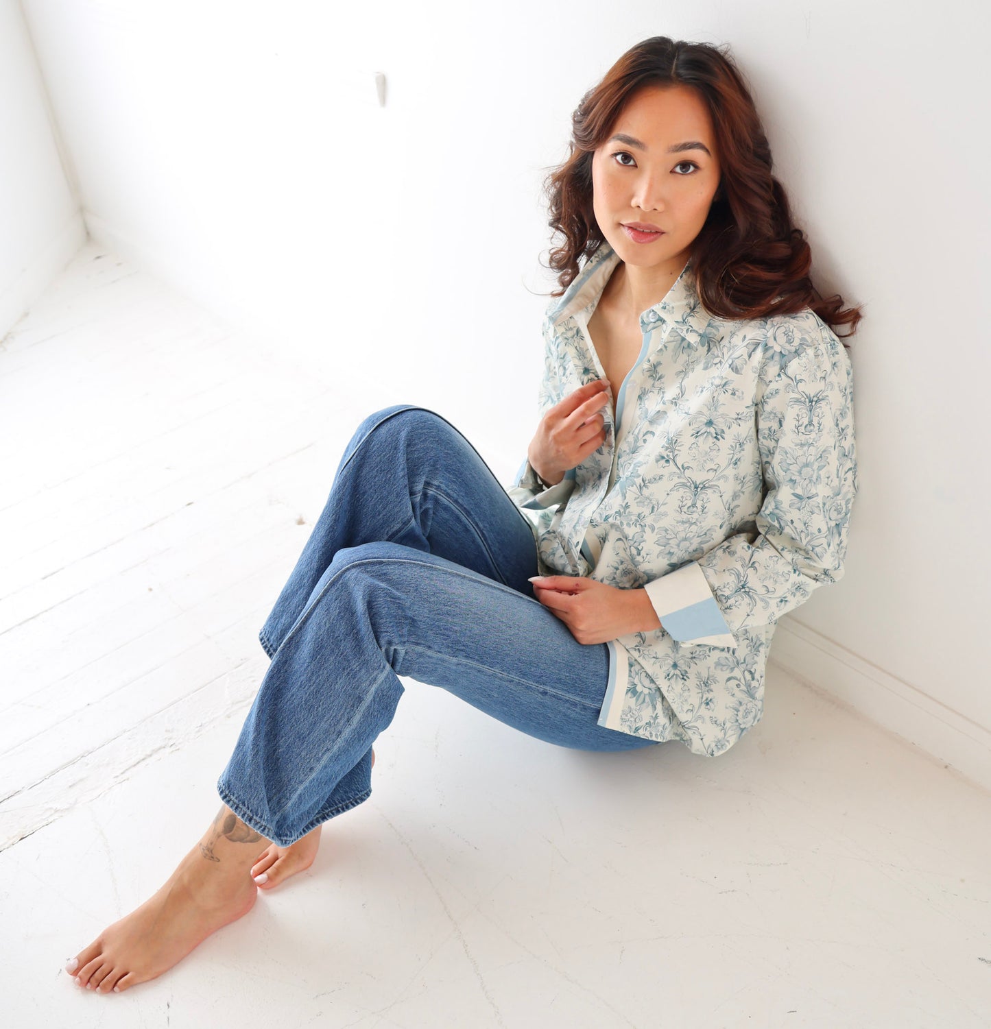 Woman sitting on a white floor wearing a floral shirt and blue jeans.