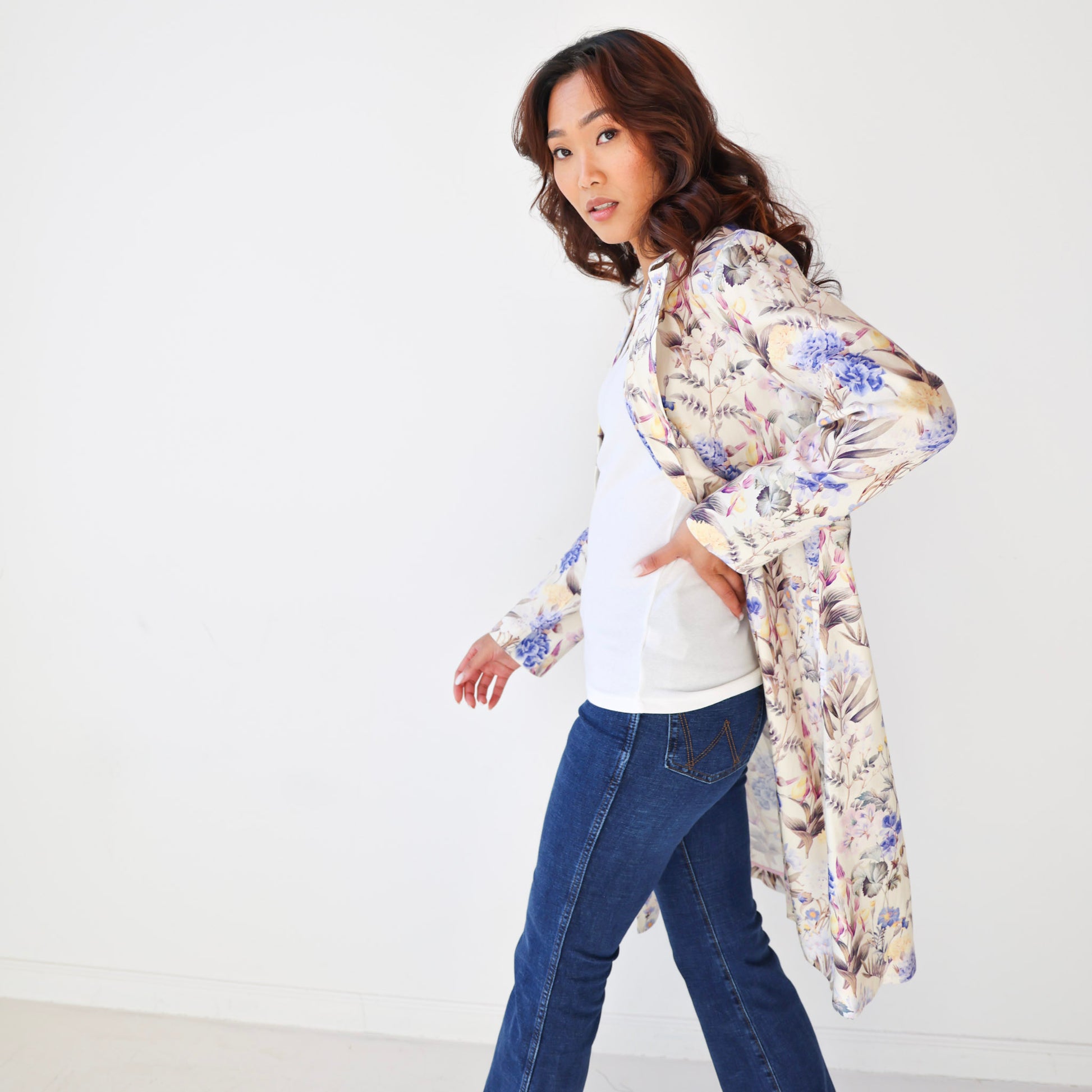 Woman wearing a floral kimono-style jacket over a white shirt and blue jeans on a plain background