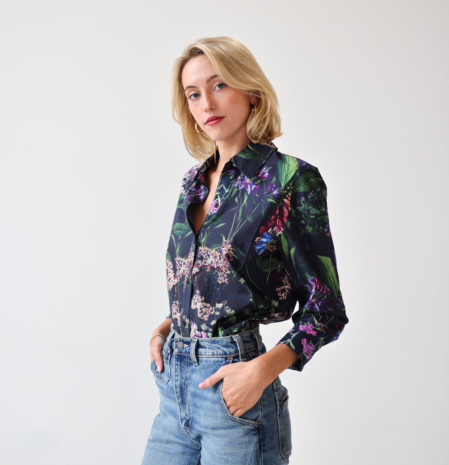 A woman with blonde hair wearing a dark floral button-down shirt and blue jeans, standing against a plain light gray background.